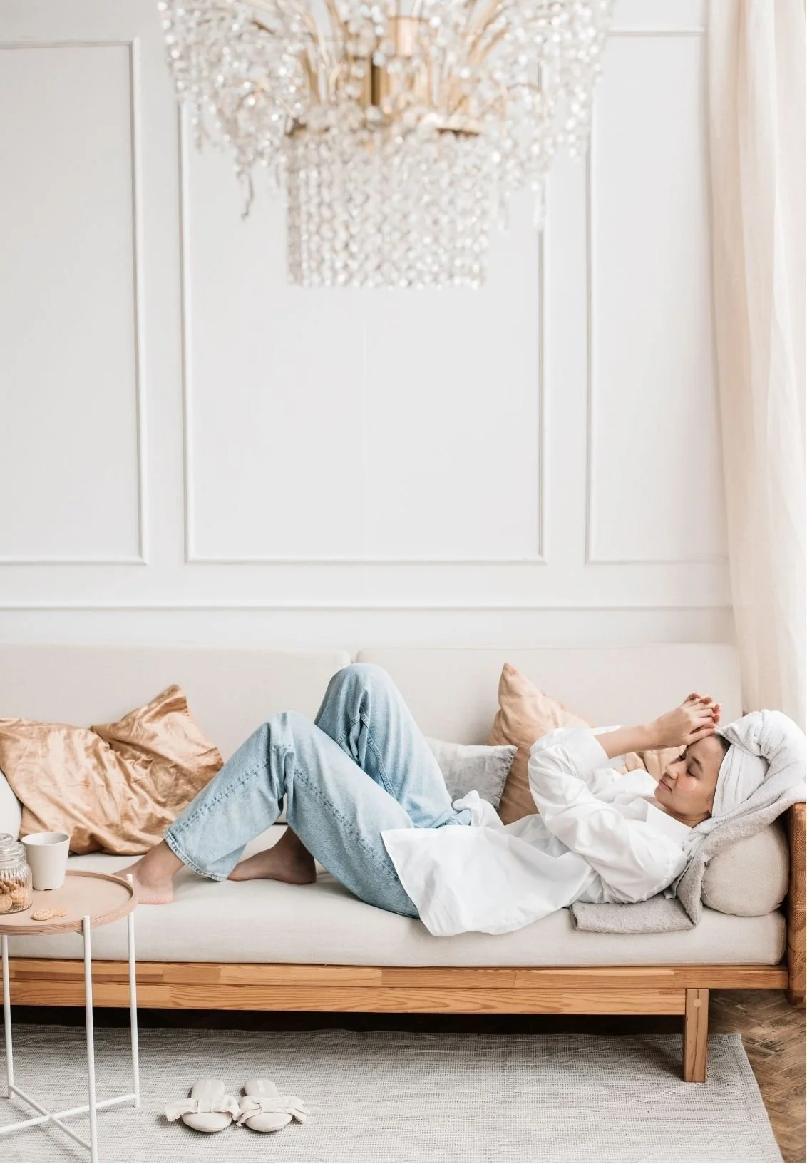 Sick woman resting on a sofa with a towel wrapped around her head, tea nearby