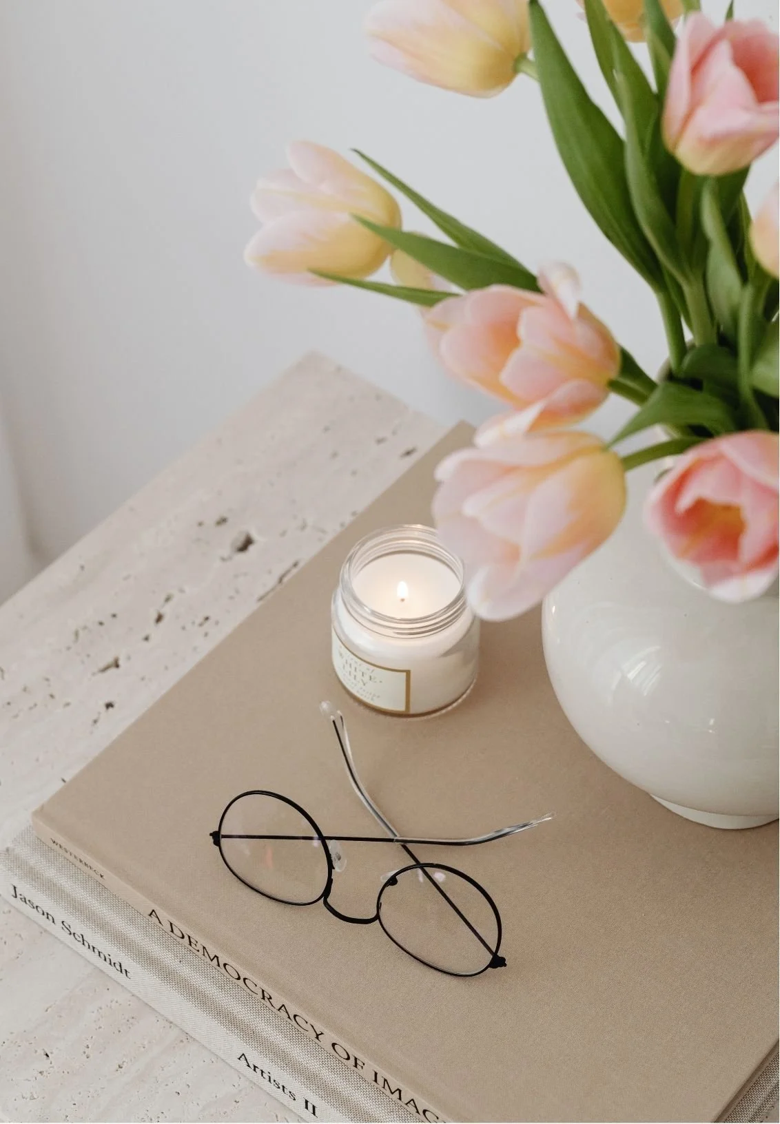 Pastel pink tulips in a white vase beside a lit candle and round eyeglasses on art books.
