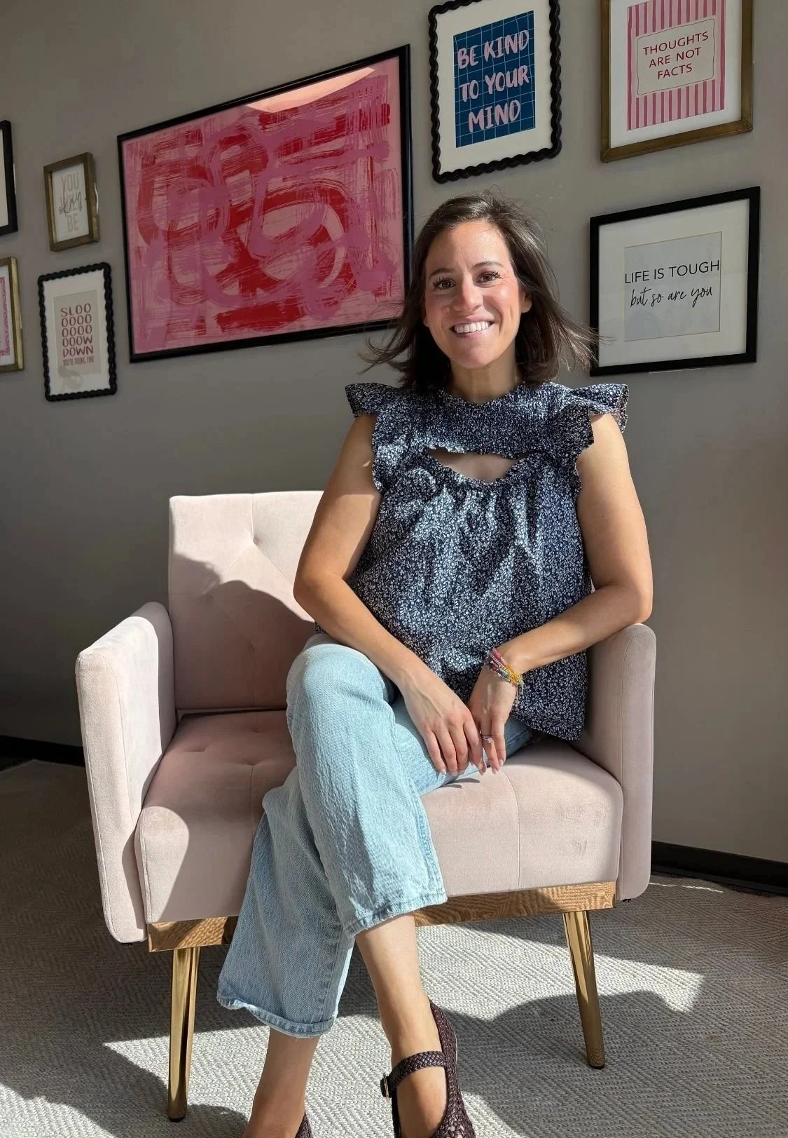 Jen Greenberg, a therapist based in North Dallas / Plano, smiling in a floral blouse and light blue jeans seated in a pink armchair beneath framed motivational wall art