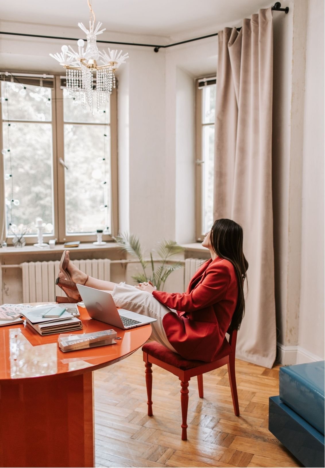 Woman in a red blazer relaxing with a laptop at a dining table in a bright room with large windows and a crystal chandelier