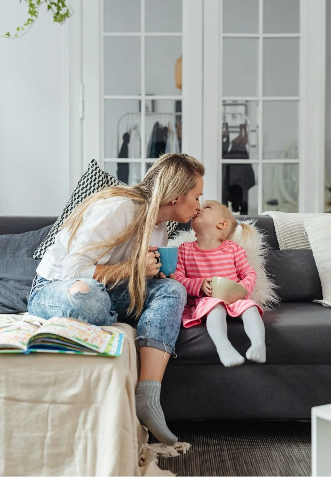 Mother kissing her young daughter on a living room sofa while holding a mug, with an open picture book nearby
