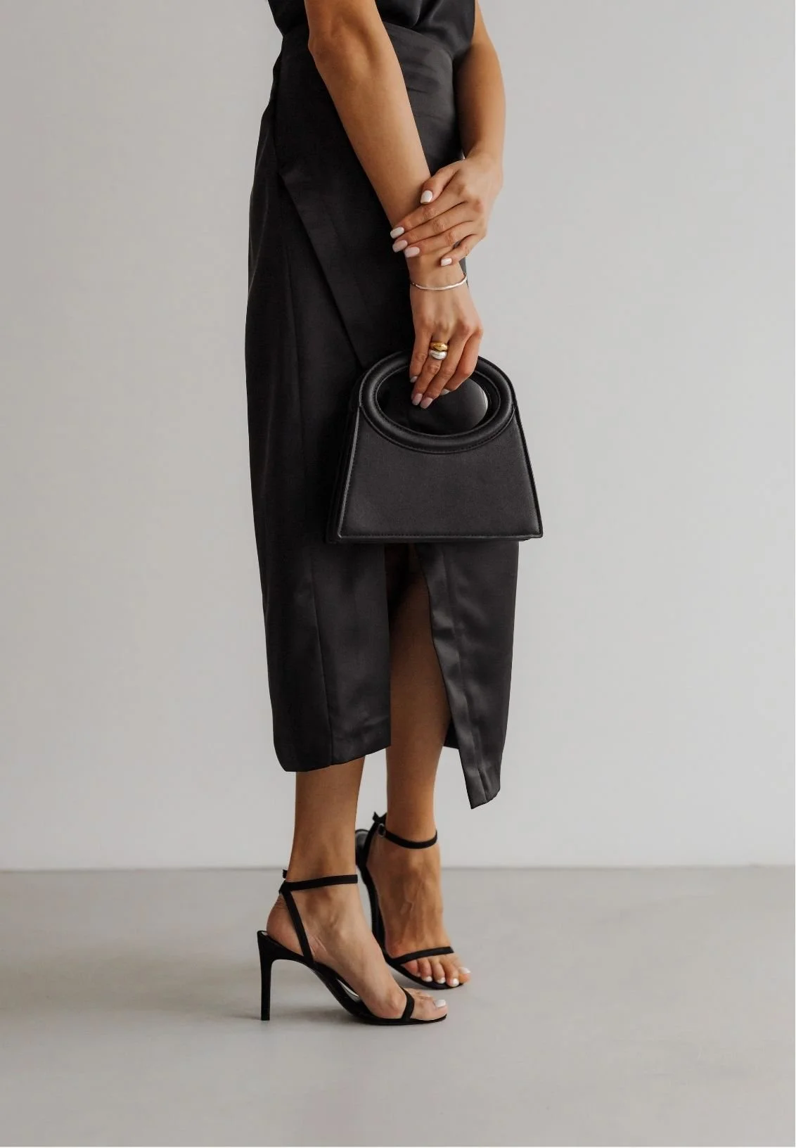 Woman in a black midi dress holding a structured black handbag and wearing black strappy high heels.