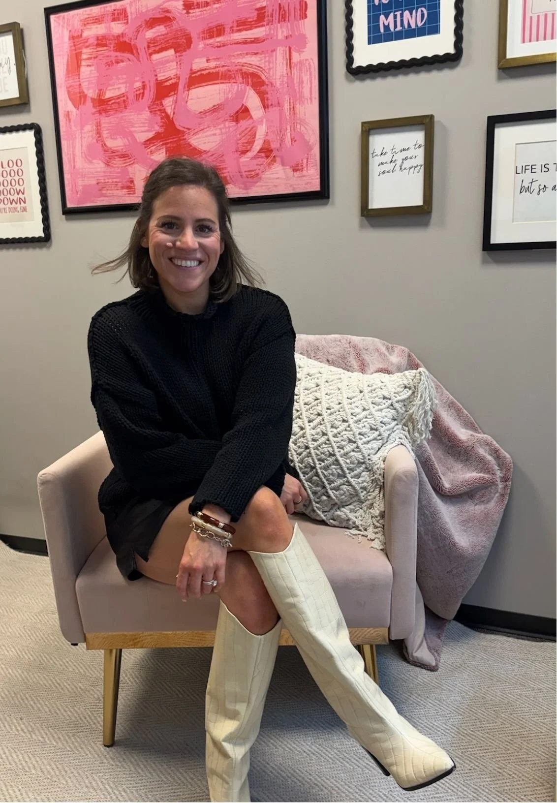 Jen Greenberg, a therapist based in North Dallas / Plano, smiling in a black sweater and white knee-high boots sitting in a pink armchair beneath colorful framed artwork