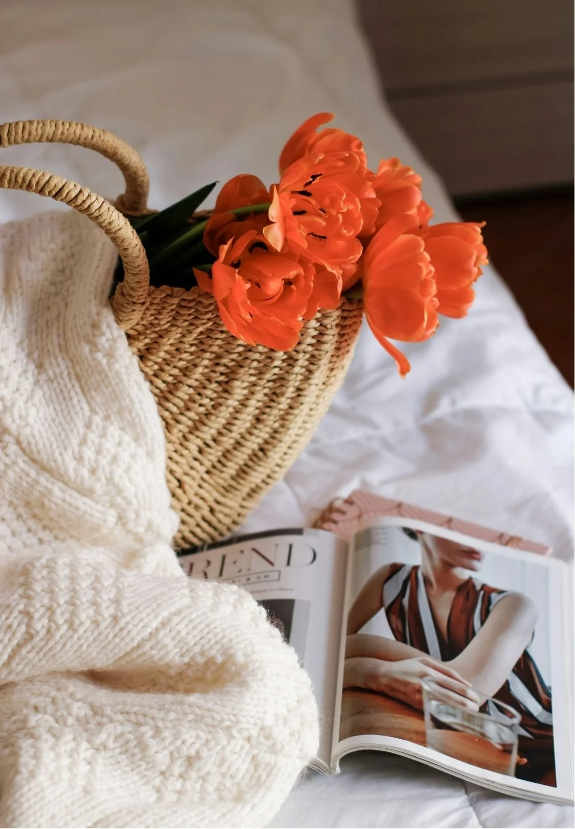 Orange tulips in a woven basket beside a cream knit blanket and open fashion magazine on a bed