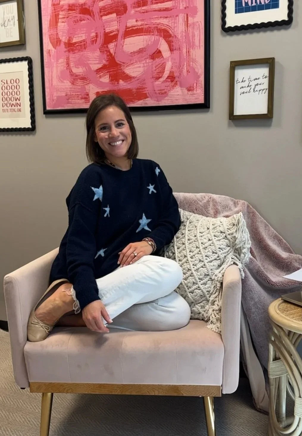 Jen Greenberg, a therapist based in North Dallas / Plano, smiling and sitting cross-legged in a blush armchair beside a textured pillow and throw blanket, surrounded by framed wall art.