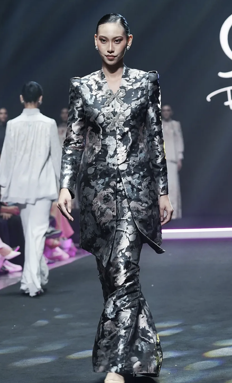 A female model walks down a runway during a fashion show, wearing a floral patterned traditional clothing with brooches on the front.