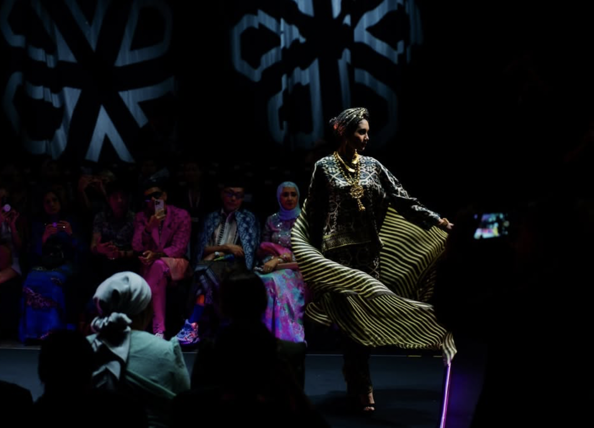 A woman in traditional patterned clothing and jewelry walks the runway, illuminated by spotlight with an audience in the background.