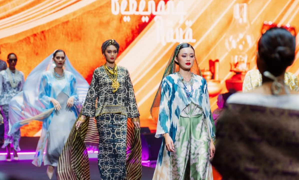 Models walking on a runway showcasing colorful traditional and modern clothing during a fashion show, with a vibrant orange background.