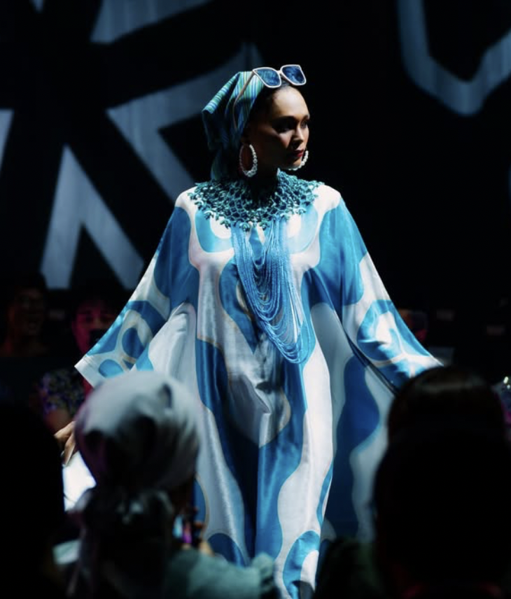 Model walking on runway, wearing a blue and white patterned flowing dress, large earrings, headwrap with sunglasses, and layered neck jewelry, at warnaraya 2026 fashion show.