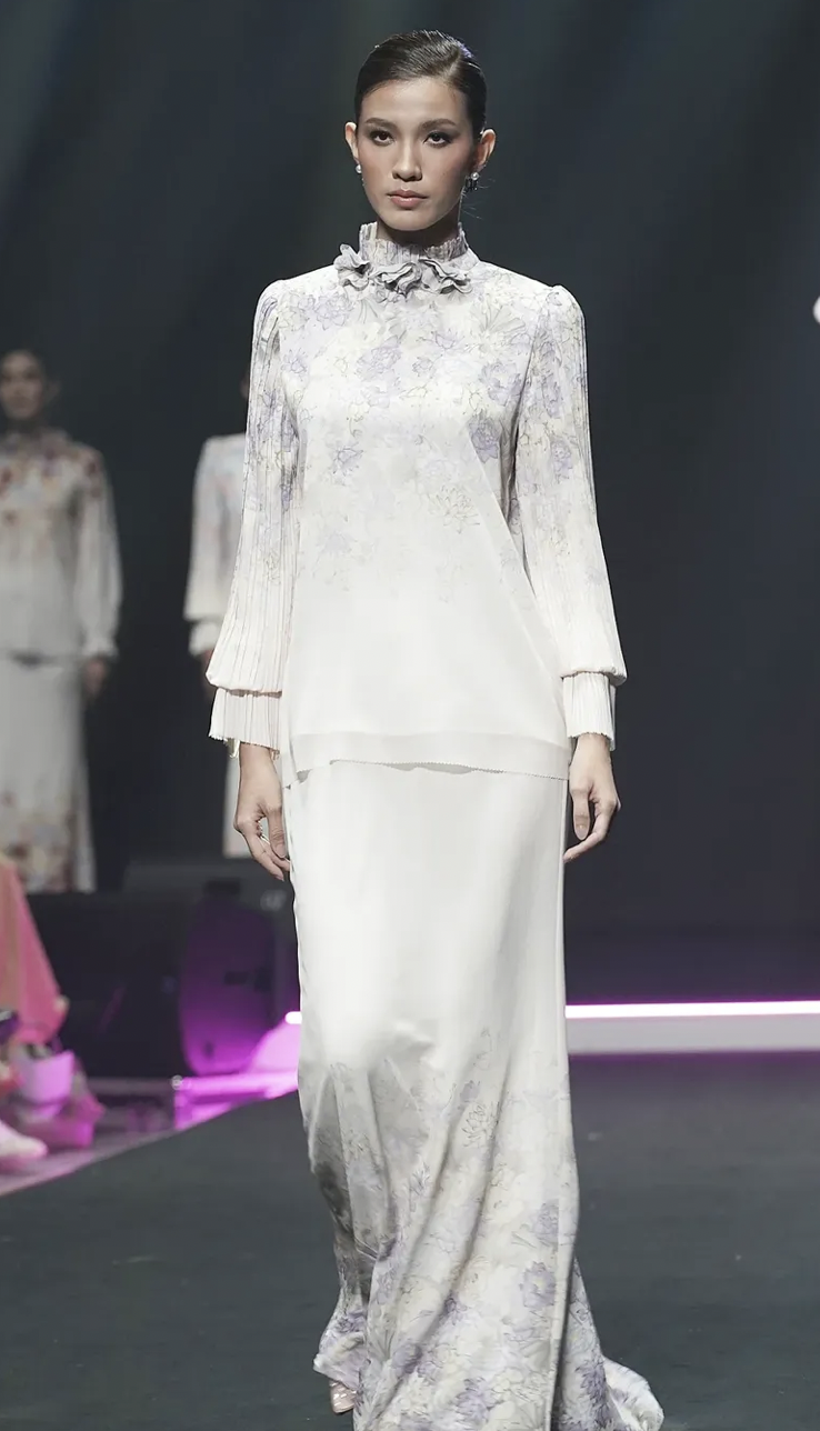 Model walking down the runway in a traditional Malay clothing, high neckline with ruffle detail, and a full-length skirt during a fashion show.