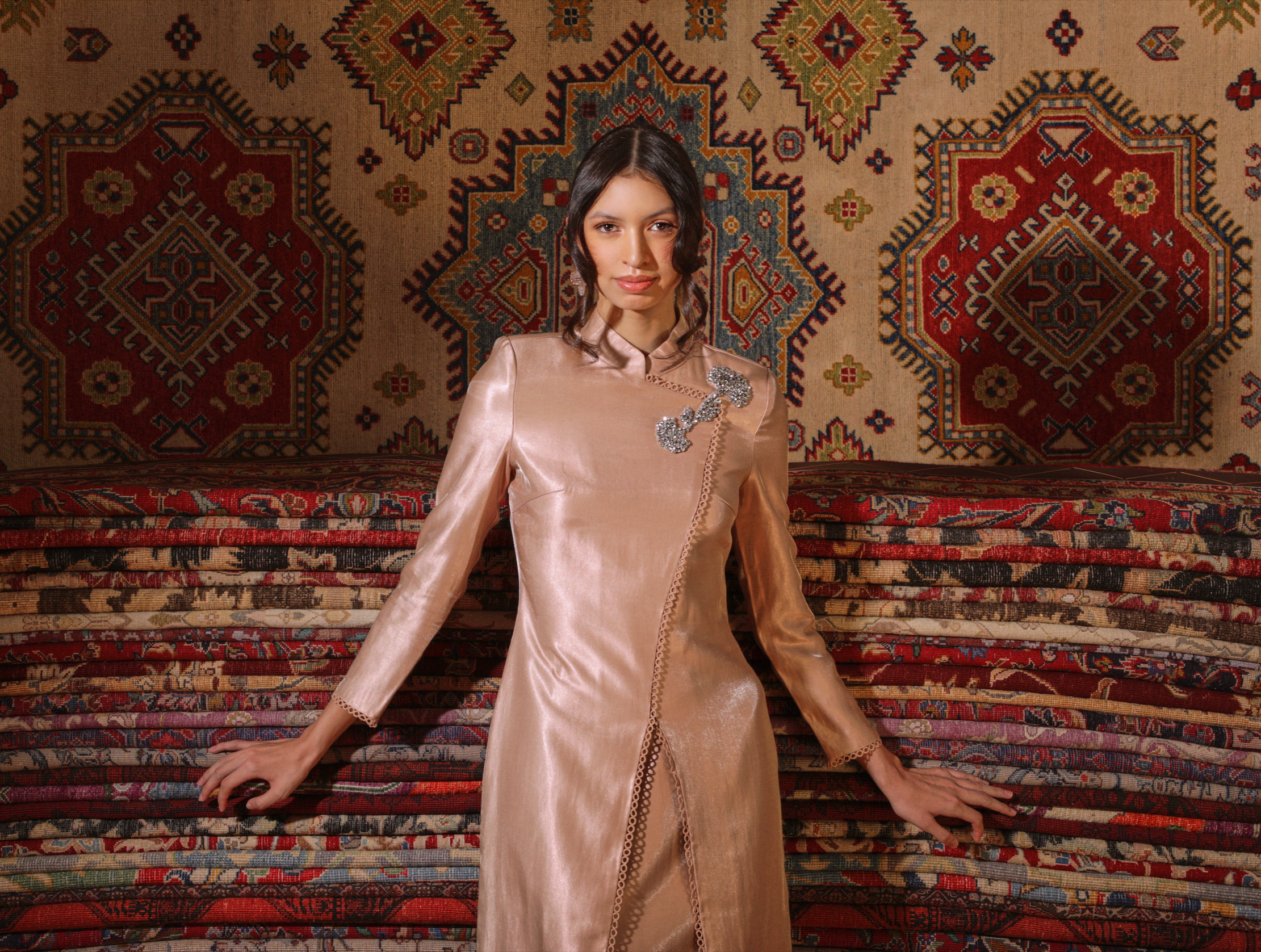 A woman with dark hair and light skin wearing a shiny pink satin traditional Malay clothing with embellishments. She is posing in front of a wall covered with a large, colorful patterned rug with geometric designs and floral motifs.