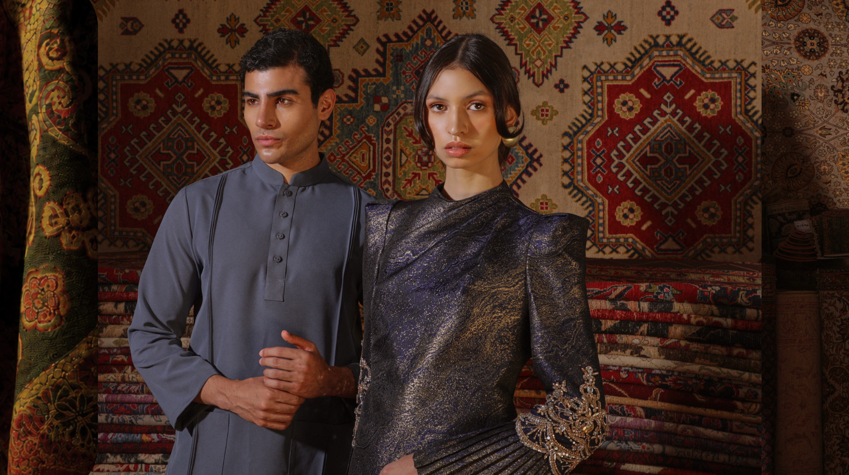 A man and a woman in traditional malay clothing posing in front of a wall with colorful, intricate rug patterns.
