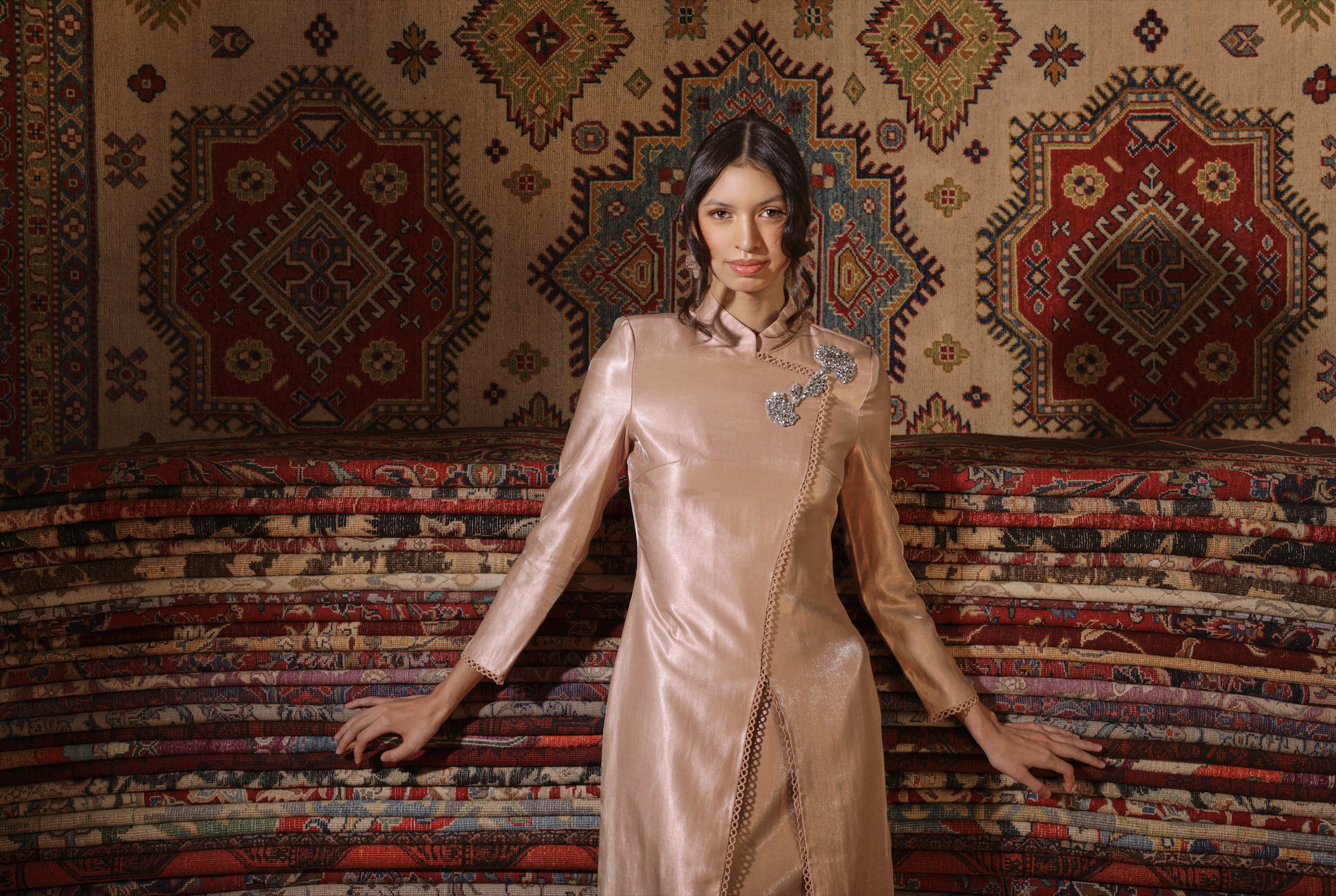 A woman with dark hair and light skin wearing a shiny pink satin traditional Malay clothing with embellishments. She is posing in front of a wall covered with a large, colorful patterned rug with geometric designs and floral motifs.