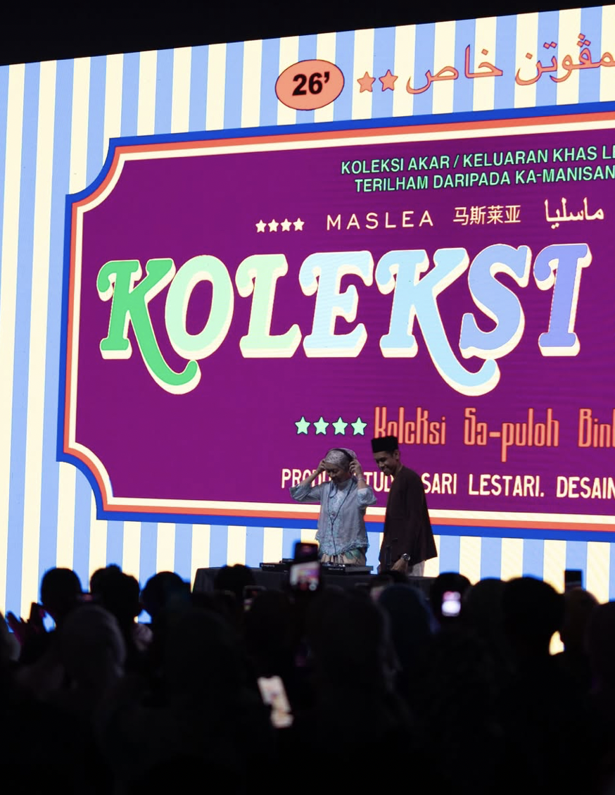 Two people standing on a stage in front of a large colorful screen with event information, with an audience watching and taking photos.