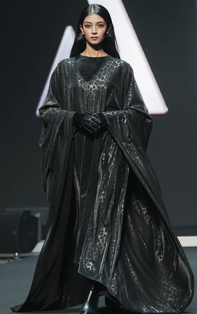 A young woman with long black hair and a serious expression is walking on a runway during a fashion show. She is wearing a shiny, black, flowing gown with a subtle leopard print pattern, black gloves, and black ankle boots.