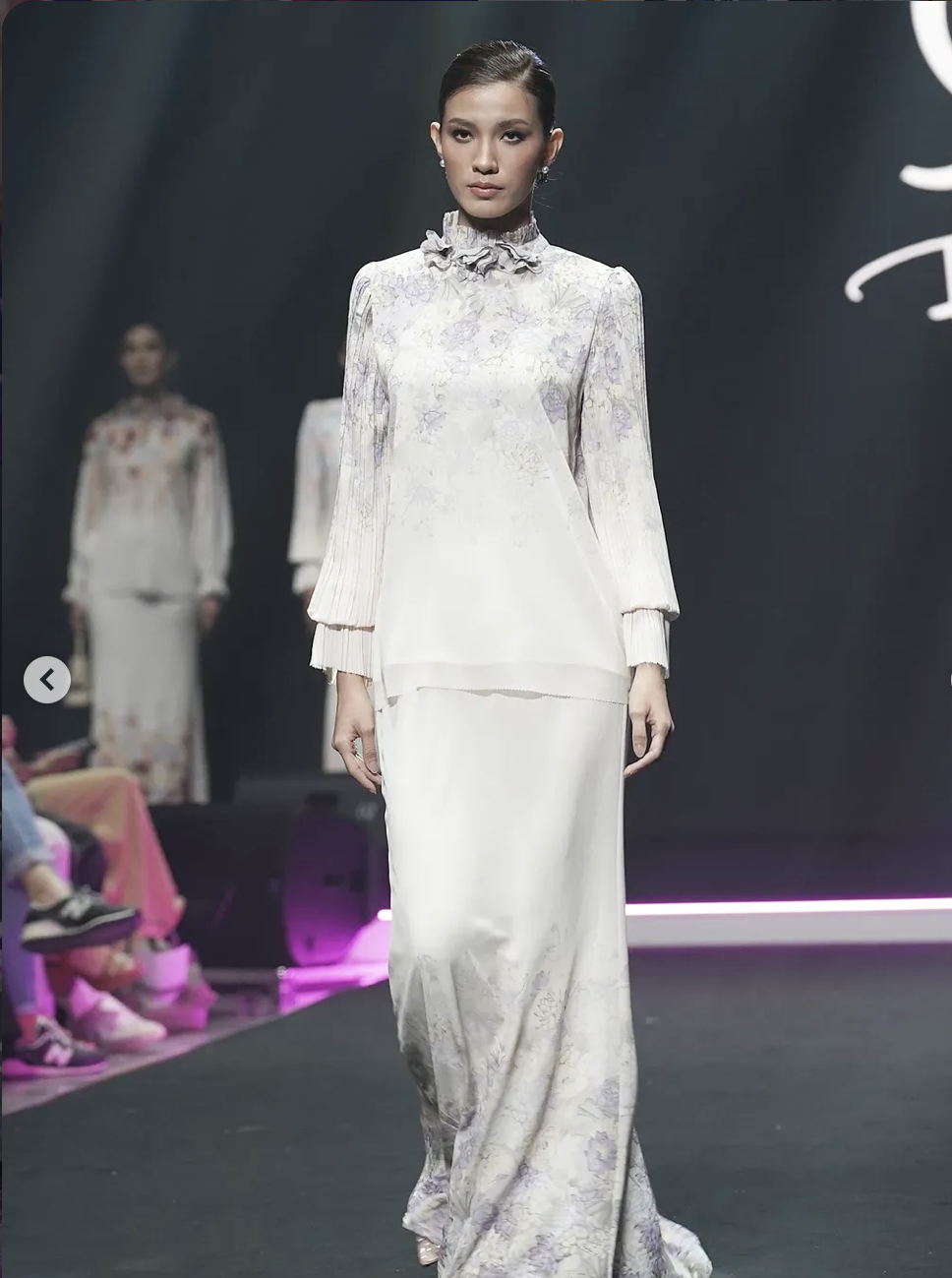 A female model walking on a runway during a fashion show, wearing a long, elegant white and purple floral traditional clothing with a high ruffled collar and long sleeves.