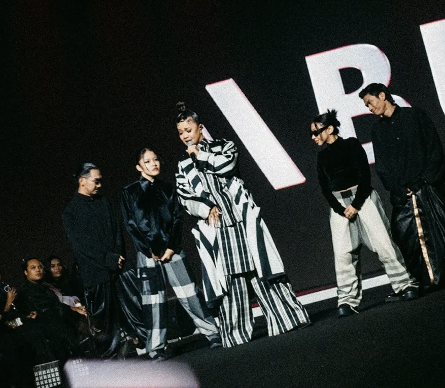Group of five individuals on stage at a fashion event, with large letterforms behind them, dressed in black and striped clothing.