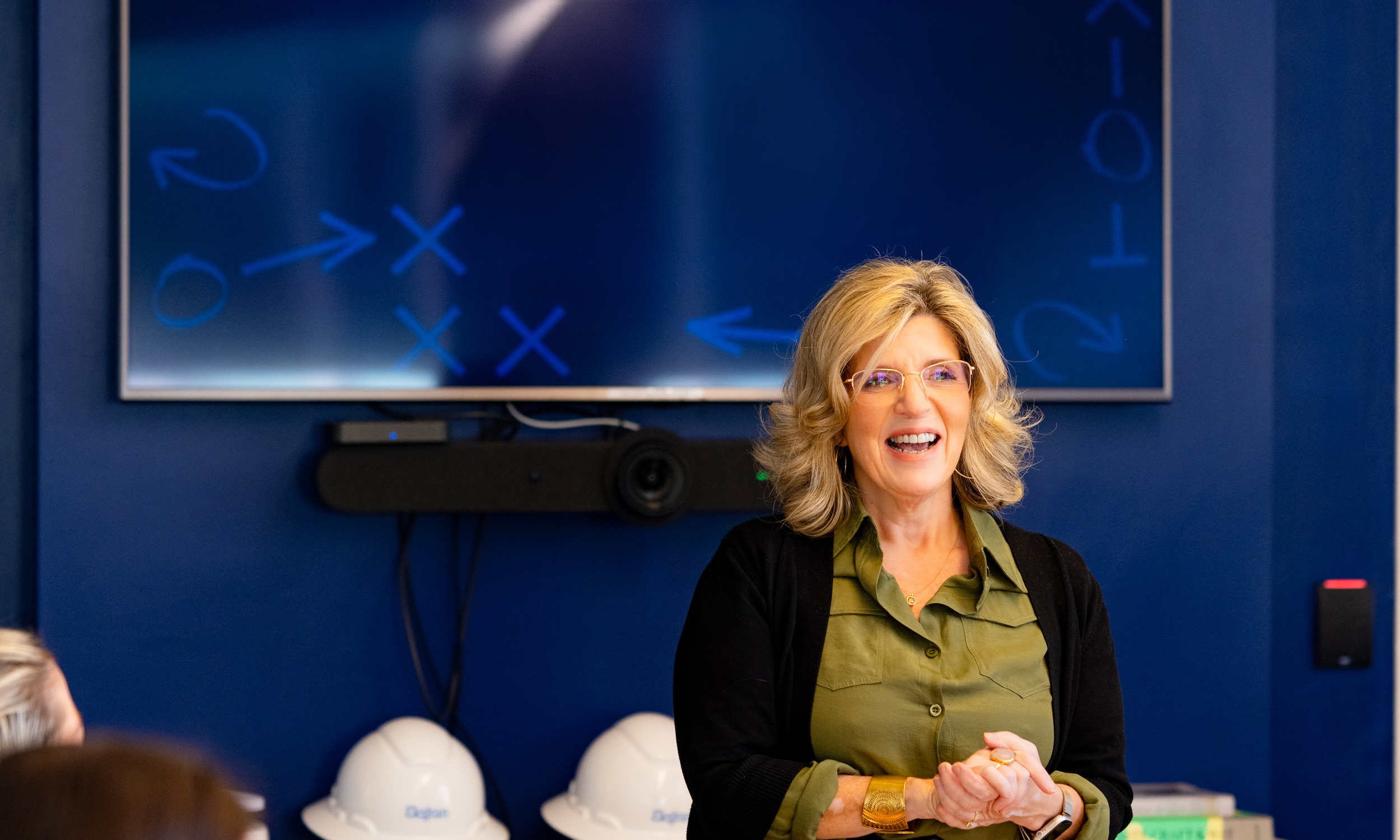 A woman smiling and standing in front of a large screen with strategy symbols, wearing glasses, a green shirt, and a black cardigan, in a conference room.