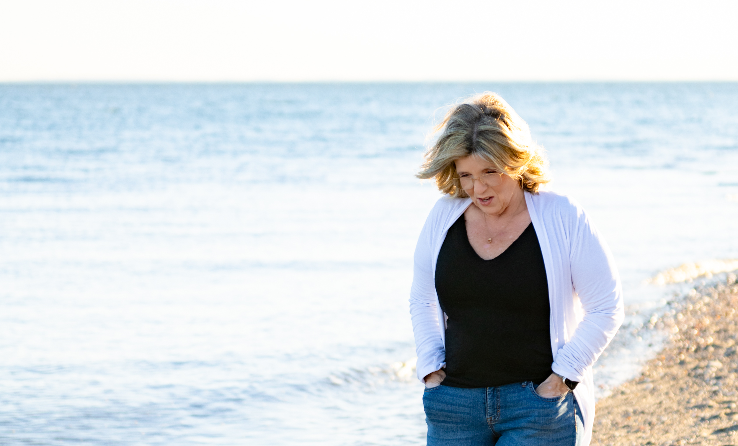 A woman with blonde hair and glasses walking on a beach near the water during sunset.