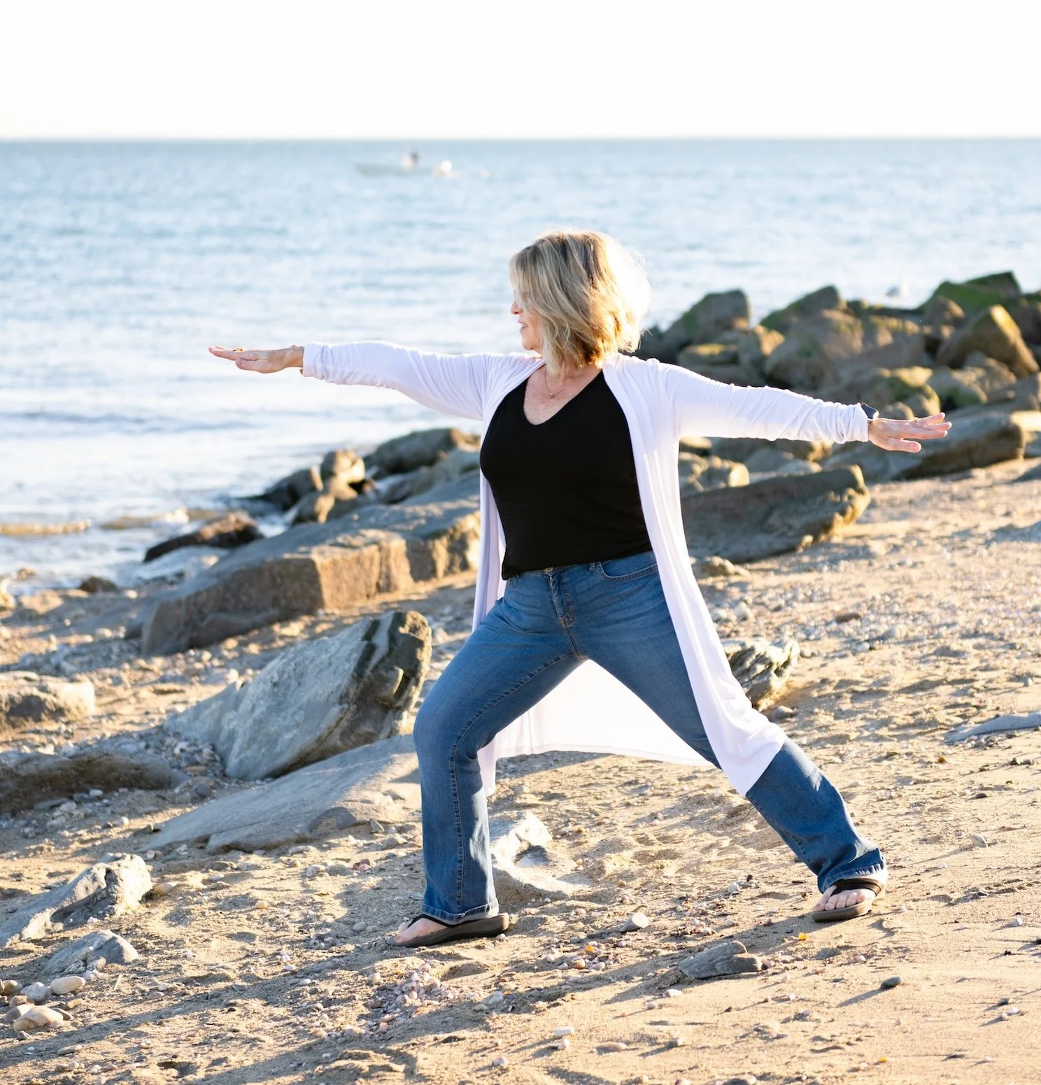 A woman practicing yoga on a beach, standing in a wide-legged stance with arms outstretched, facing the ocean during sunset.