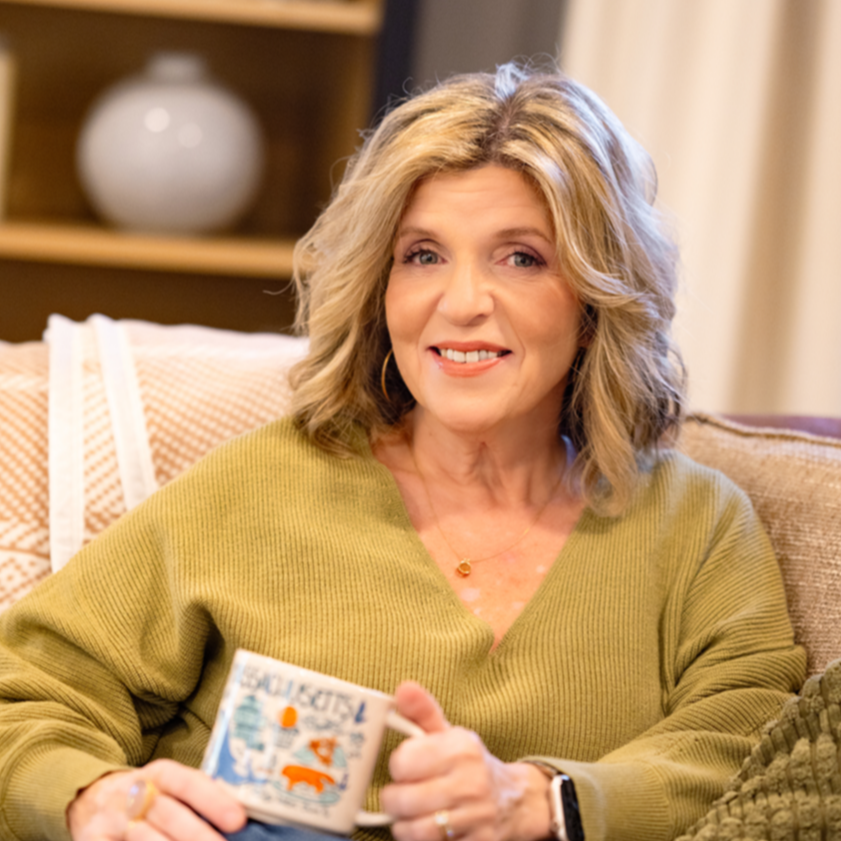 A middle-aged woman with blonde, curly hair sitting on a beige couch, holding a mug, smiling, in a cozy living room setting.