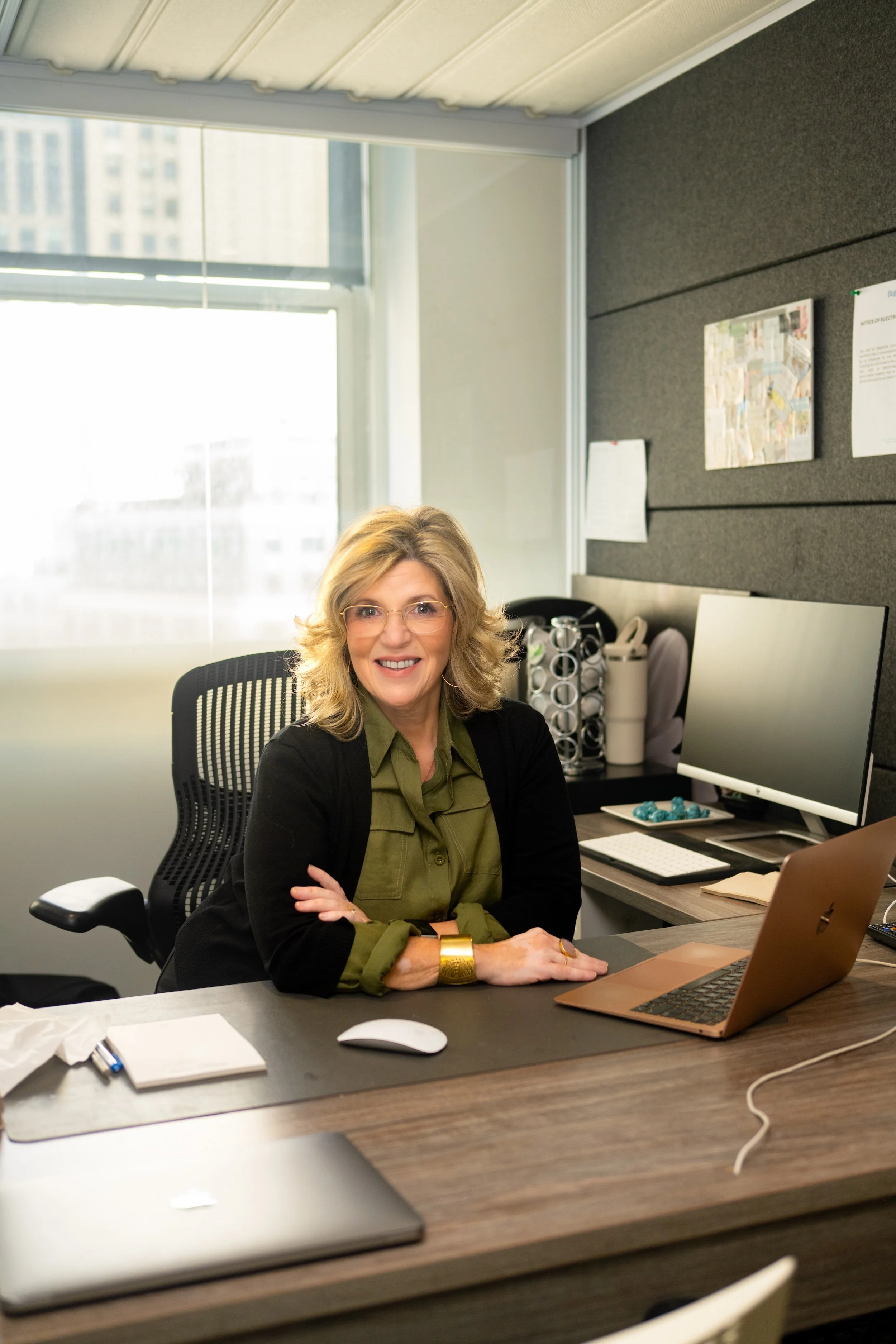 A woman with blonde hair, glasses, and wearing a green shirt and black cardigan, sitting at an office desk with her arms crossed. The desk has a laptop, mouse, and notepad. Behind her is a window with city buildings outside, and the office wall has bulletin boards and office supplies.