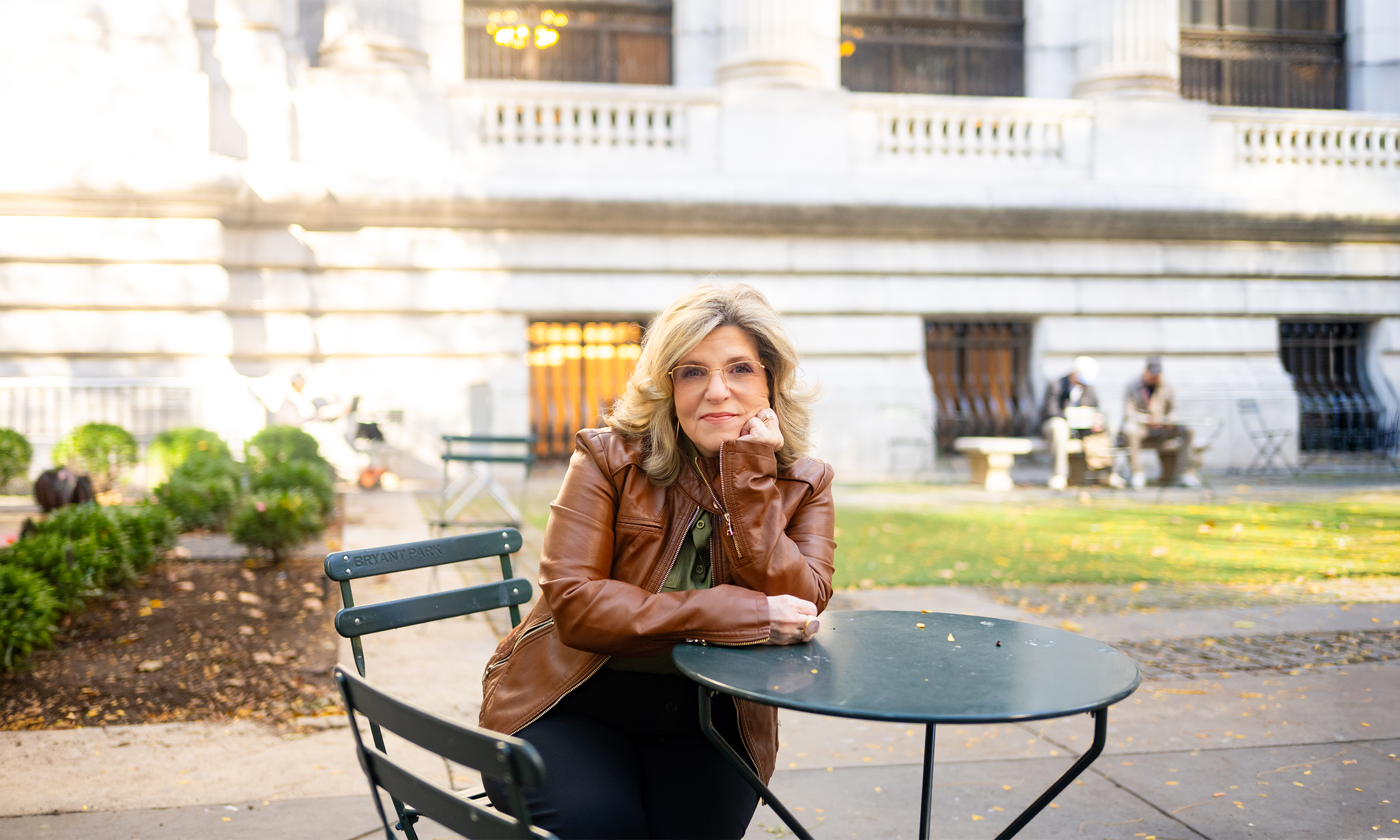 A woman with blonde hair, wearing glasses and a brown leather jacket, sitting at a dark-colored round outdoor table in a park, with a historic building with columns in the background.