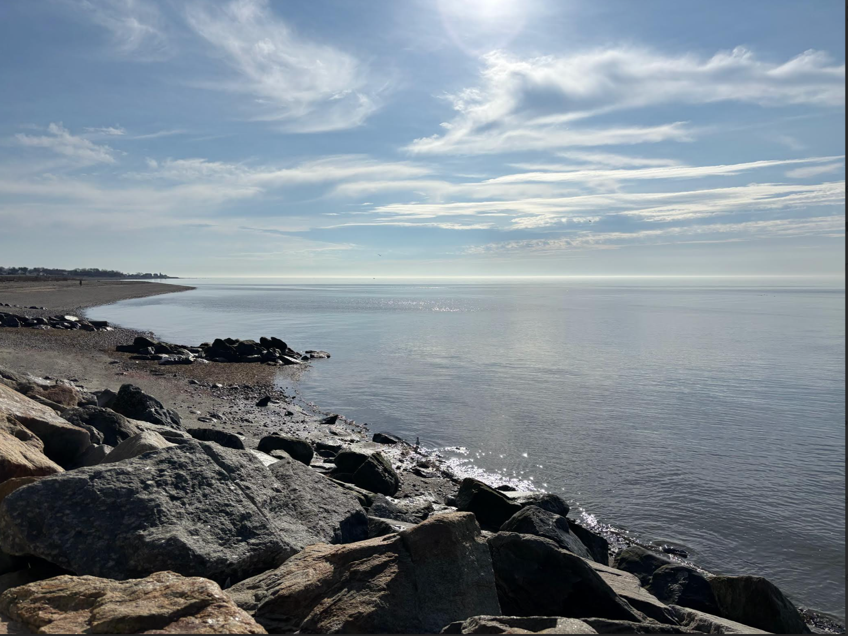 Calm ocean water along a rocky shoreline on a partly cloudy day, with light reflected on the water surface and a distant horizon.
