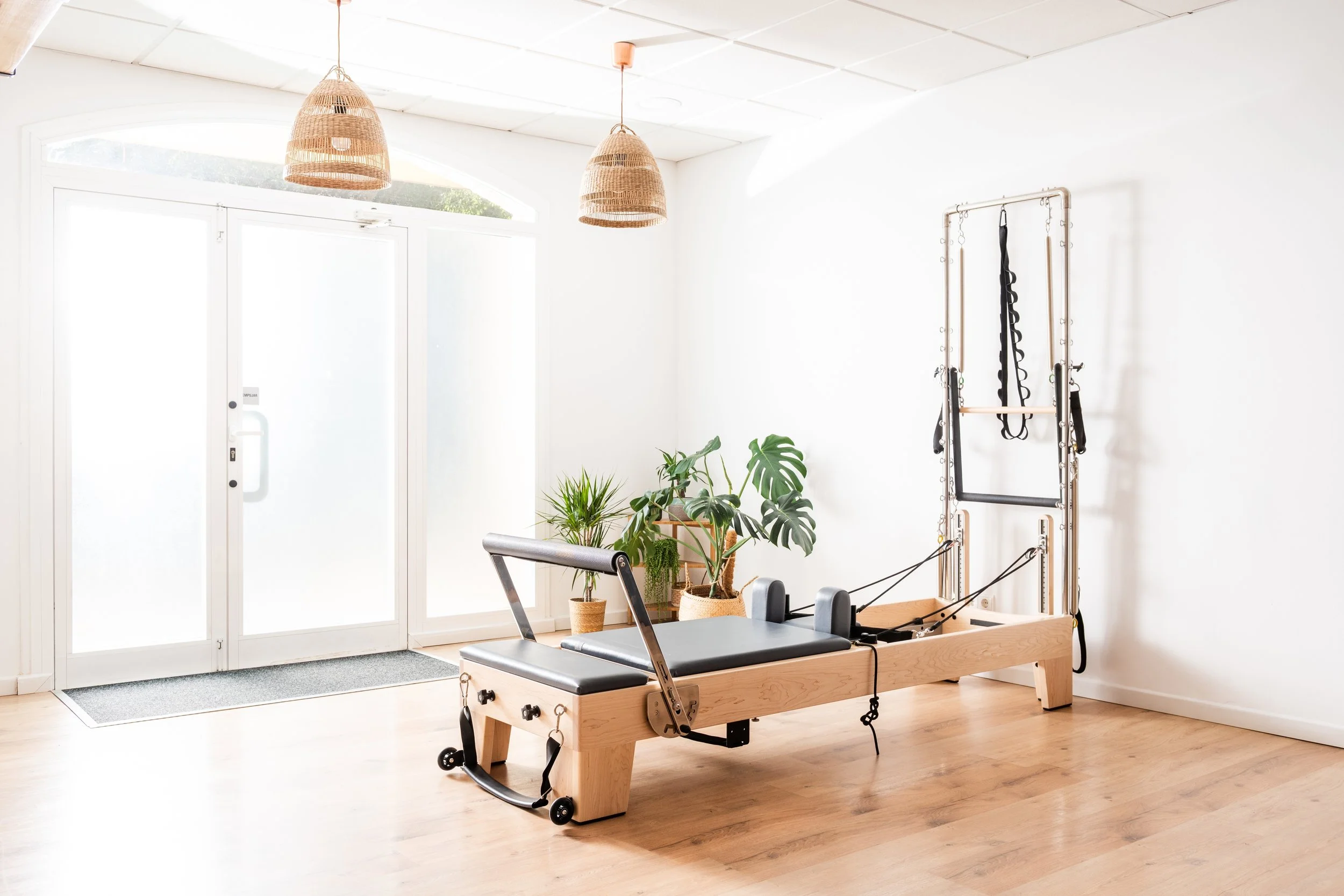 A Pilates reformer machine in a bright, minimalist fitness studio with large windows, wooden flooring, and indoor plants.