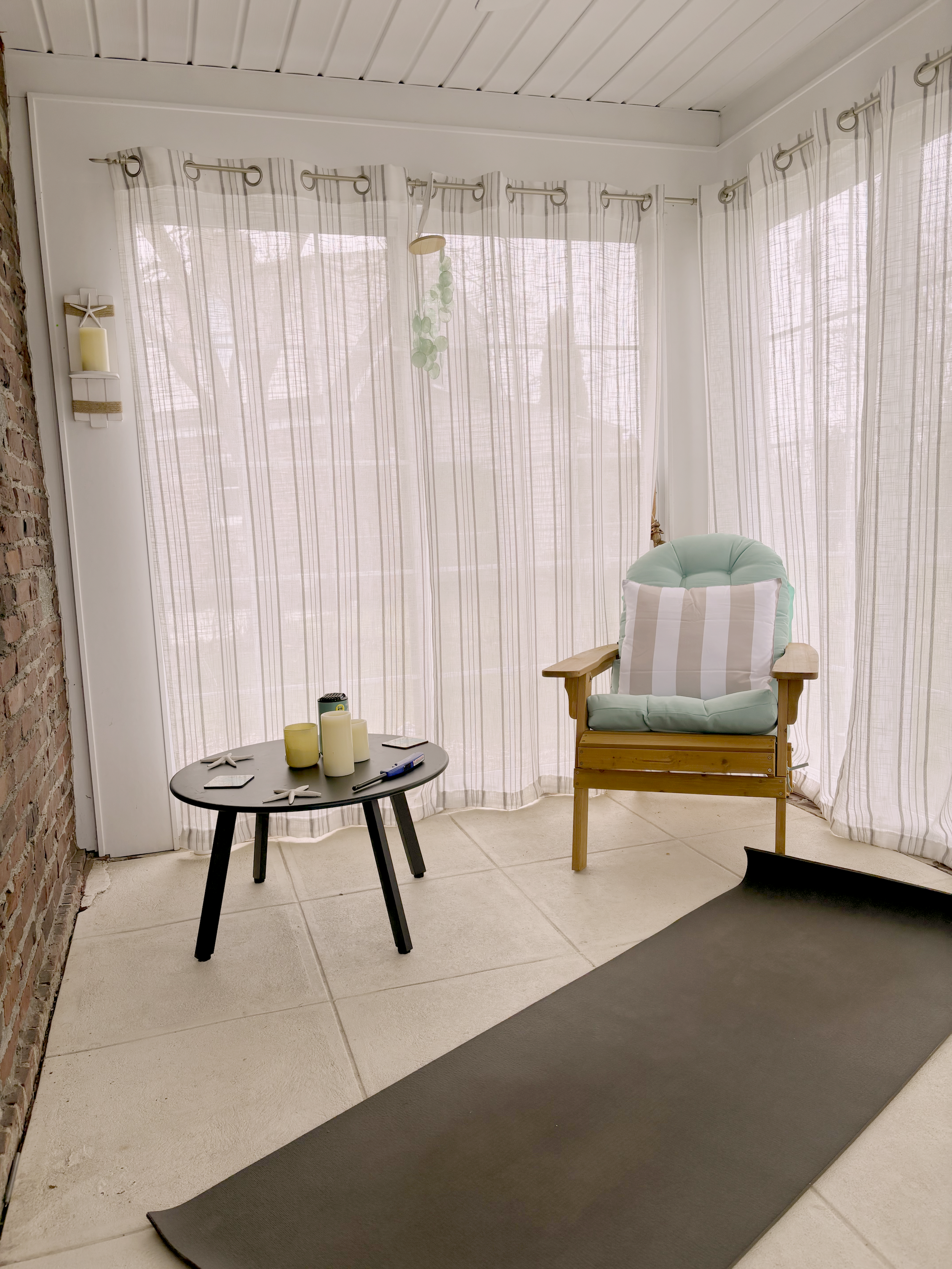 A cozy indoor space with sheer curtains on large windows, a wooden armchair with a green cushion and striped pillow, a small round black table with candles and starfish decorations, and a dark yoga mat on a tiled floor, with brick wall on one side and white paneled ceiling.