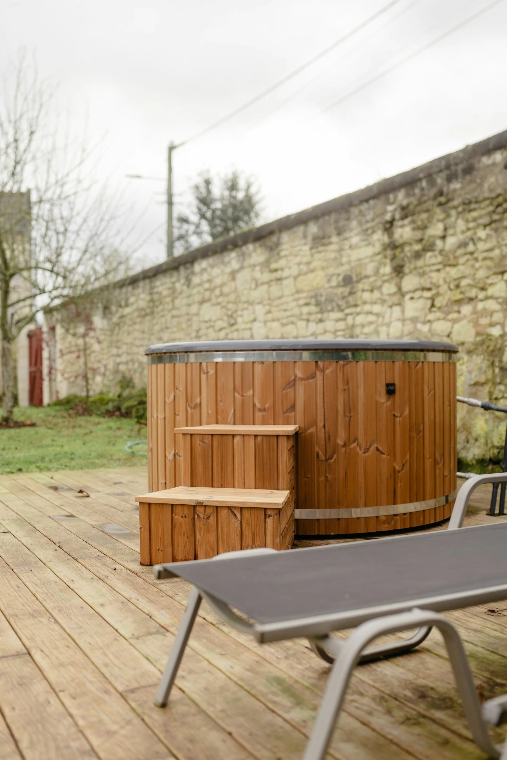 An outdoor wooden hot tub with a small set of wooden steps leading up to it, set on a wooden deck. In the background, there is a stone wall, some trees, and a cloudy sky.