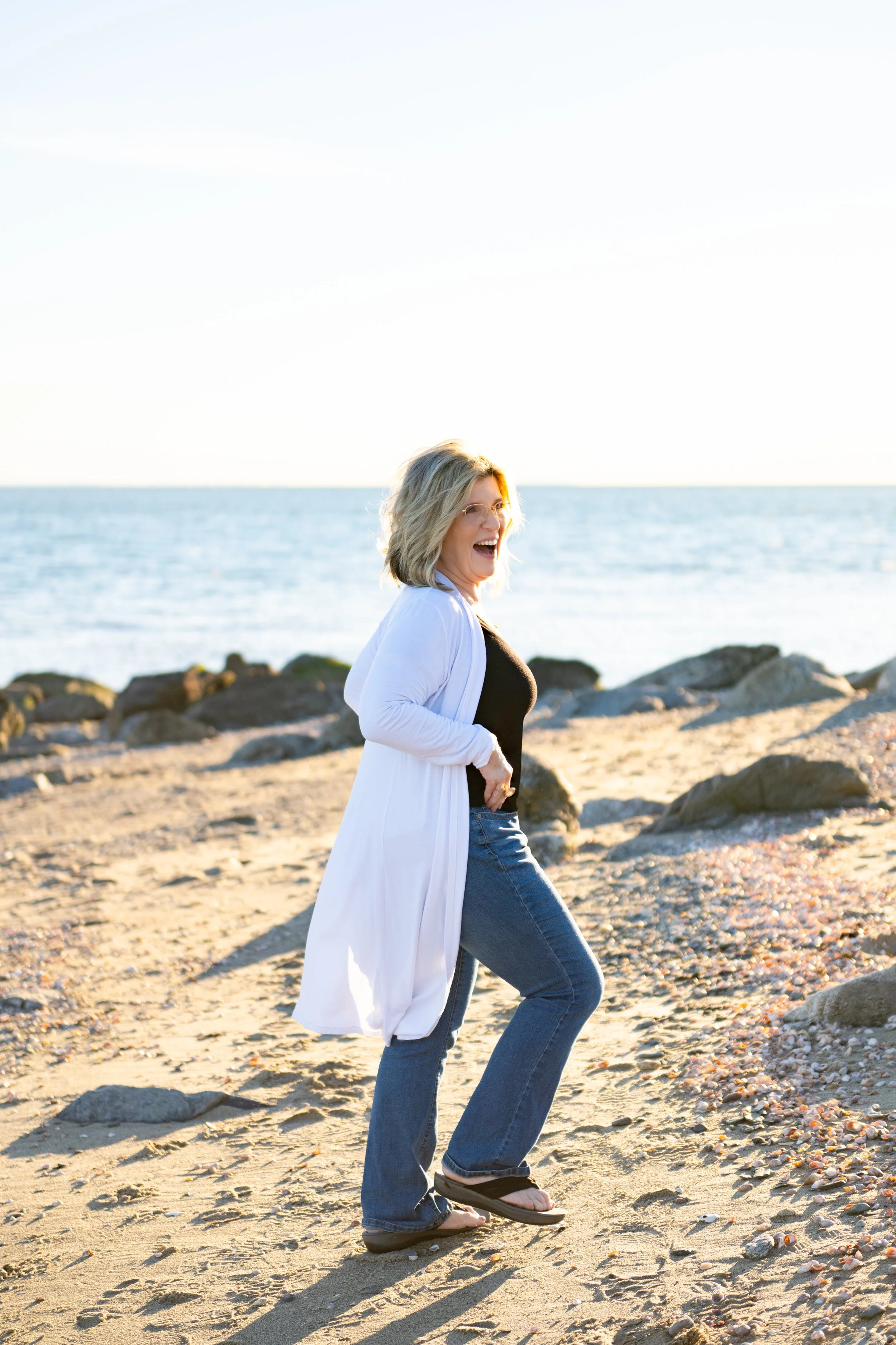 A woman wearing glasses, a white cardigan, black top, and jeans laughing while walking on a beach with rocks and pebbles near the water.
