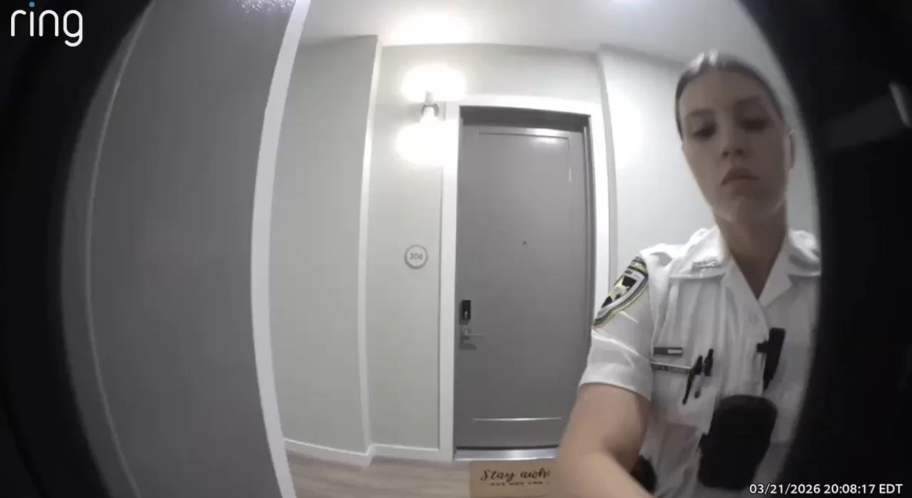 Security camera footage showing a female police officer with dark hair in a white uniform, standing in front of a gray door in an apartment or hotel corridor