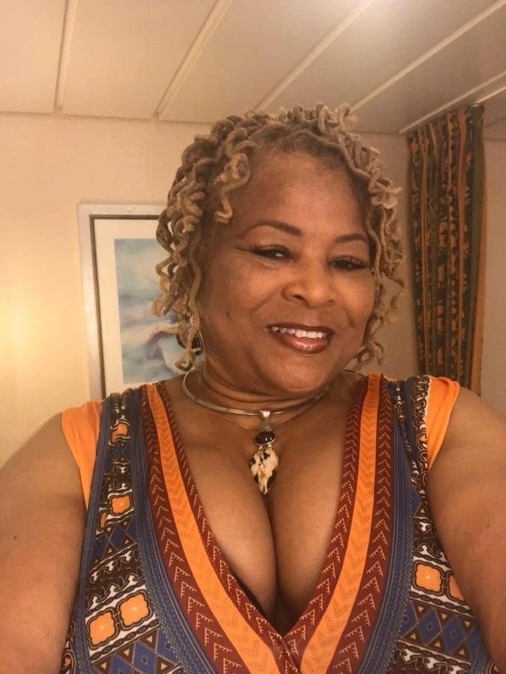 Close-up of a woman with blonde curly hair taking a selfie indoors, smiling, wearing a colorful patterned dress and a necklace.