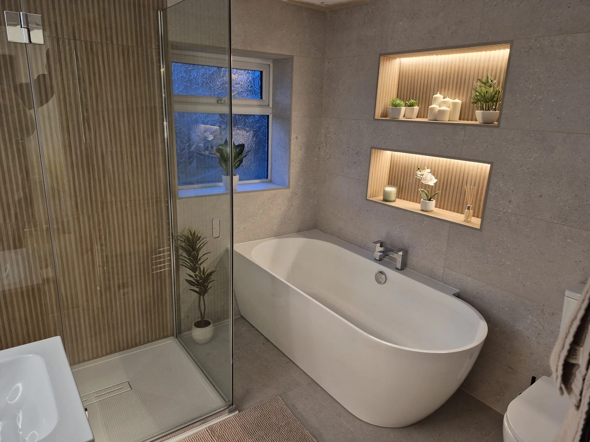 Modern bathroom with a freestanding bathtub against a wall with built-in illuminated shelves holding white candles and green plants, window with frosted glass, glass shower enclosure, and gray tiled walls.