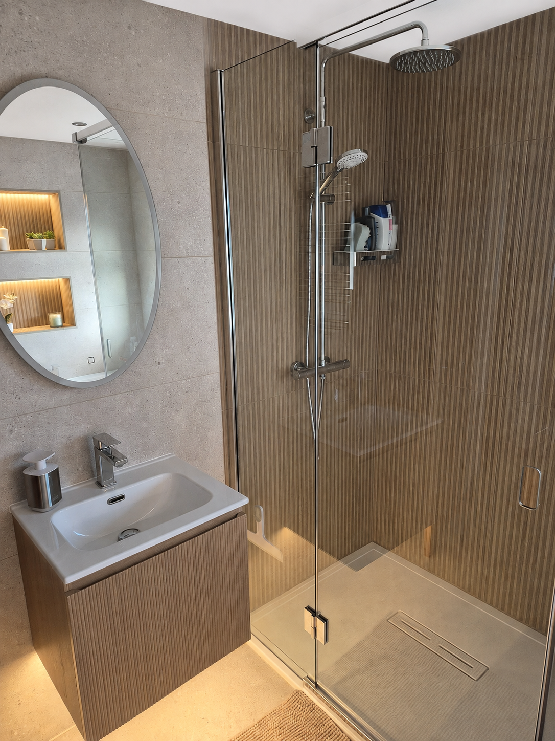 A modern bathroom with a glass-enclosed shower area, a white sink with a wooden cabinet, and a wallpapered wall behind the sink with an oval mirror.