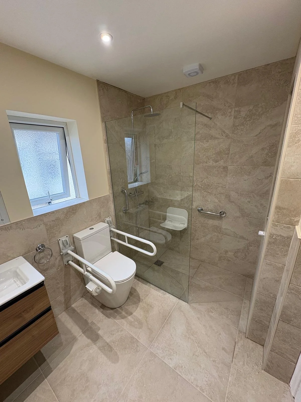 A modern accessible bathroom with a toilet, grab bars, a shower area with a glass partition, a small window, and beige tiles on the floor and walls.