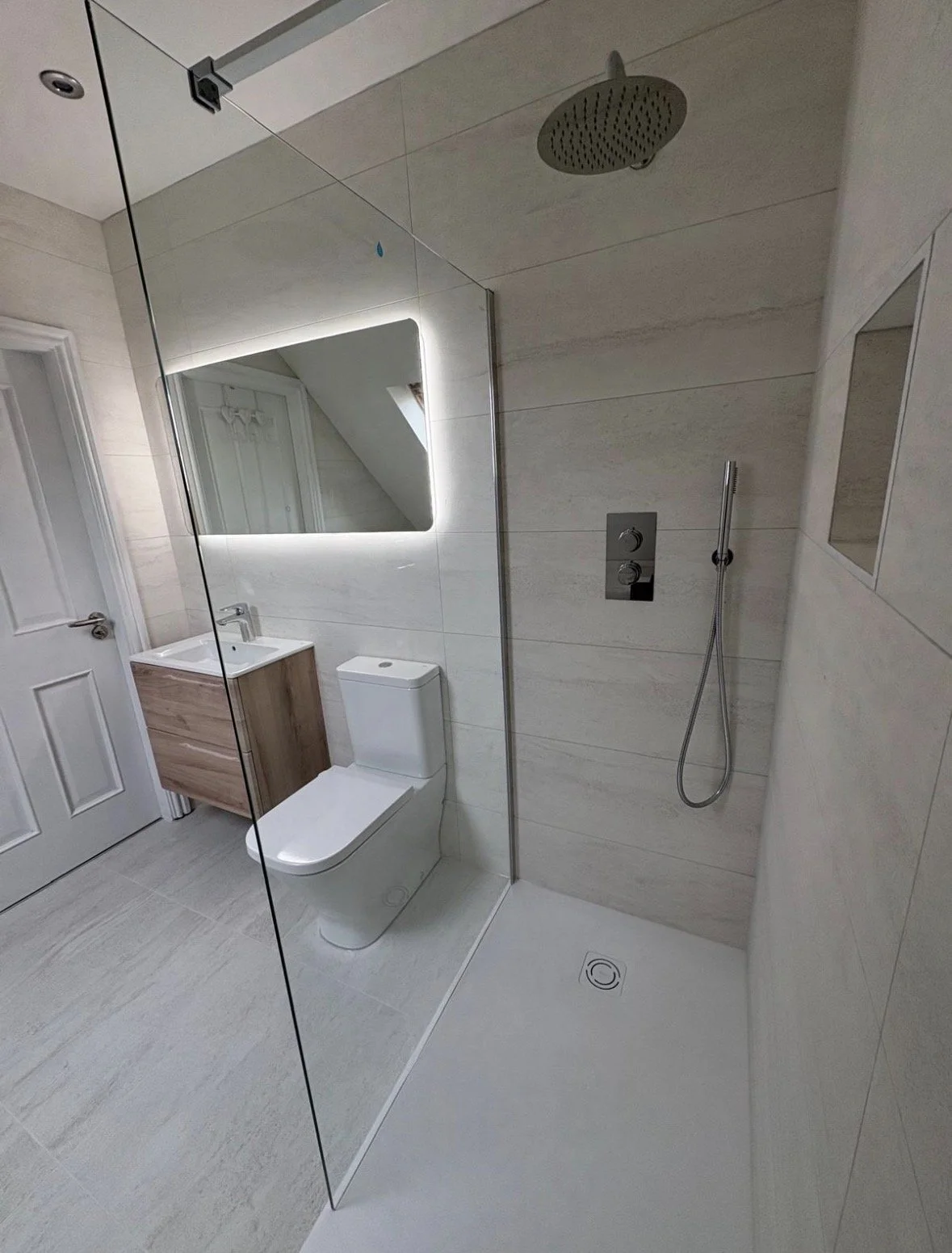 Modern bathroom with a walk-in shower, a wall-mounted mirror with LED lighting, a toilet, a small wooden vanity with a sink, and light-colored tiled walls and floor.