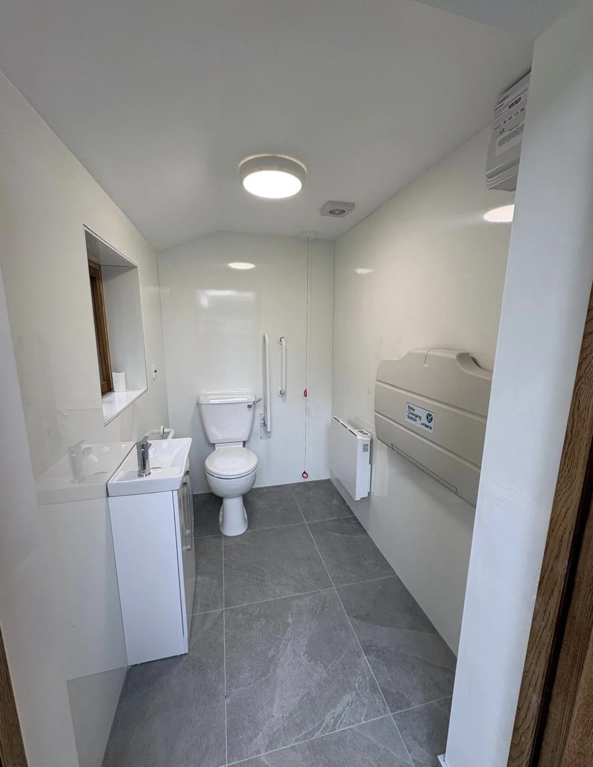 Accessible bathroom with white walls, gray tile flooring, a toilet, a small white sink, grab bars, and an automatic hand dryer.