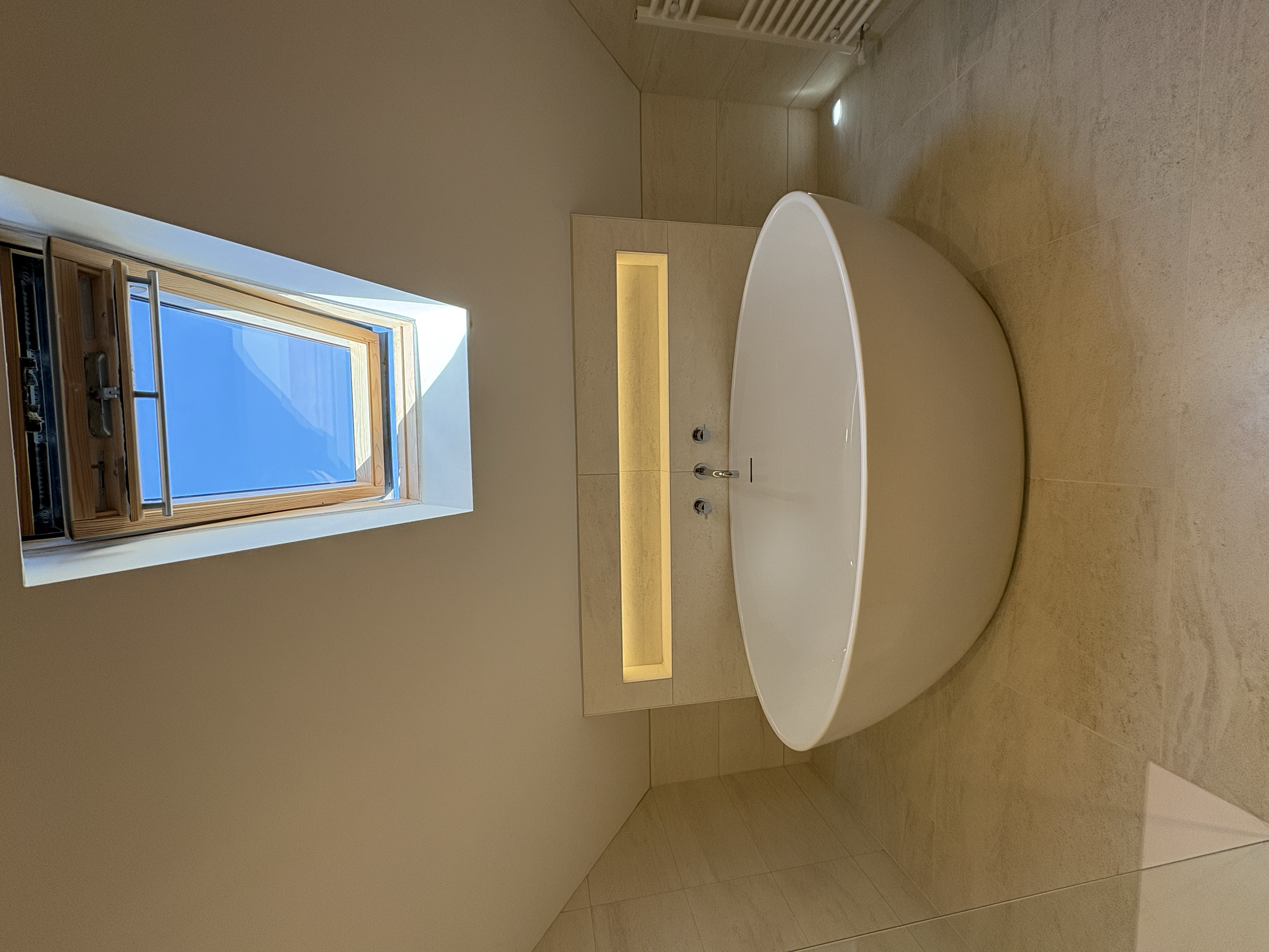 A modern bathroom with a freestanding oval bathtub, a skylight window on the ceiling, beige tiled walls, and a built-in lighted shelf above the tub.