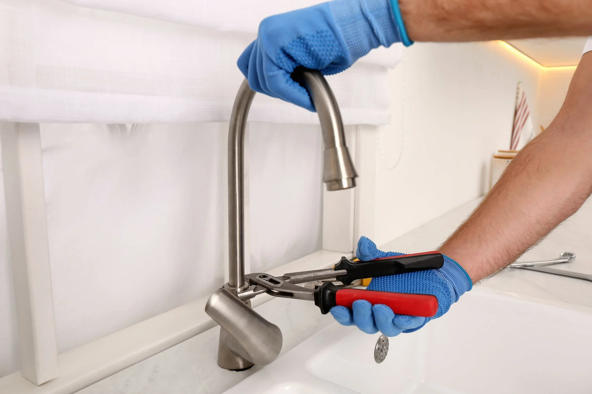 Person wearing blue gloves tightening a pipe under a white sink with pliers.