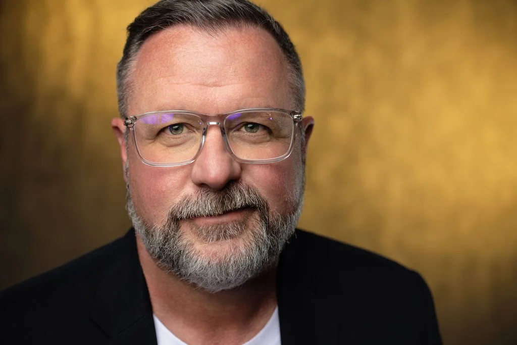Close-up portrait of a middle-aged man with gray hair, a beard, wearing glasses, in front of a gold-colored background.