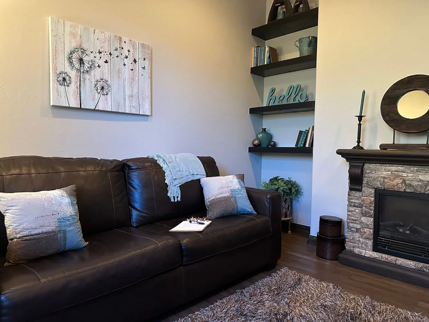 Living room with a dark brown leather sofa, a stone fireplace, a framed dandelion and butterflies wall art, and black floating shelves with books, vases, and an ornament reading 'hello'. There is also a side table, candles, and a few pillows on the sofa.