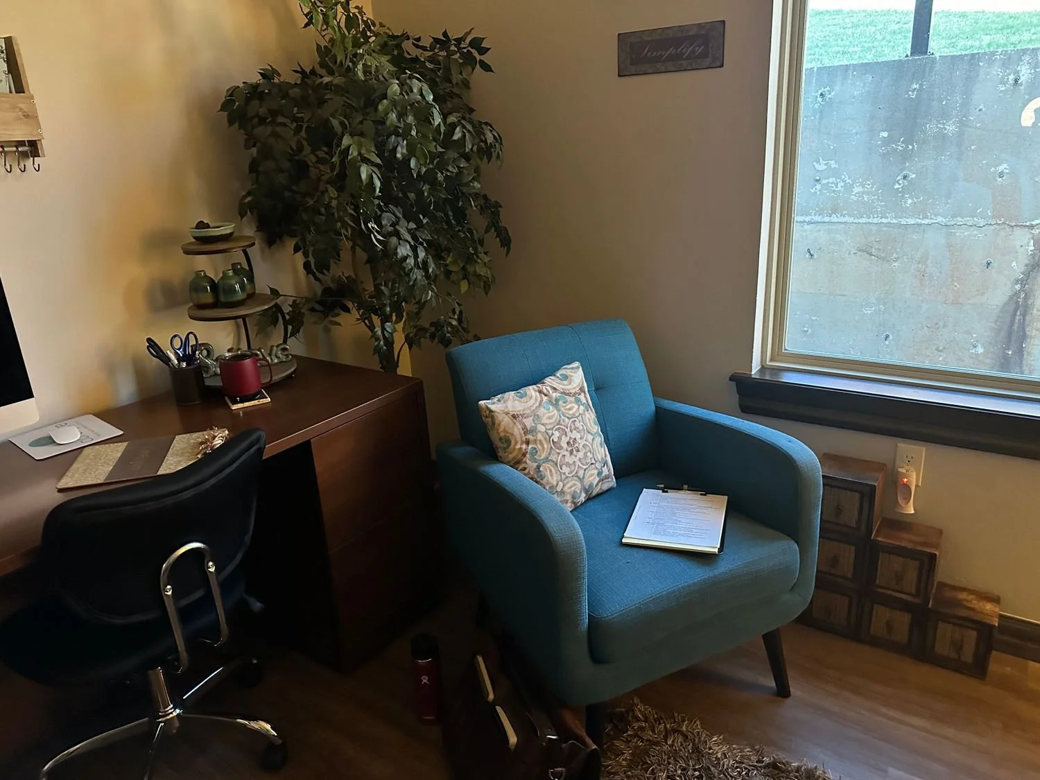 A cozy home office corner with a blue armchair holding a patterned pillow and a clipboard with papers, next to a window, a small side table, and a tall artificial plant. There is a desk with a computer monitor, a chair, and office supplies.