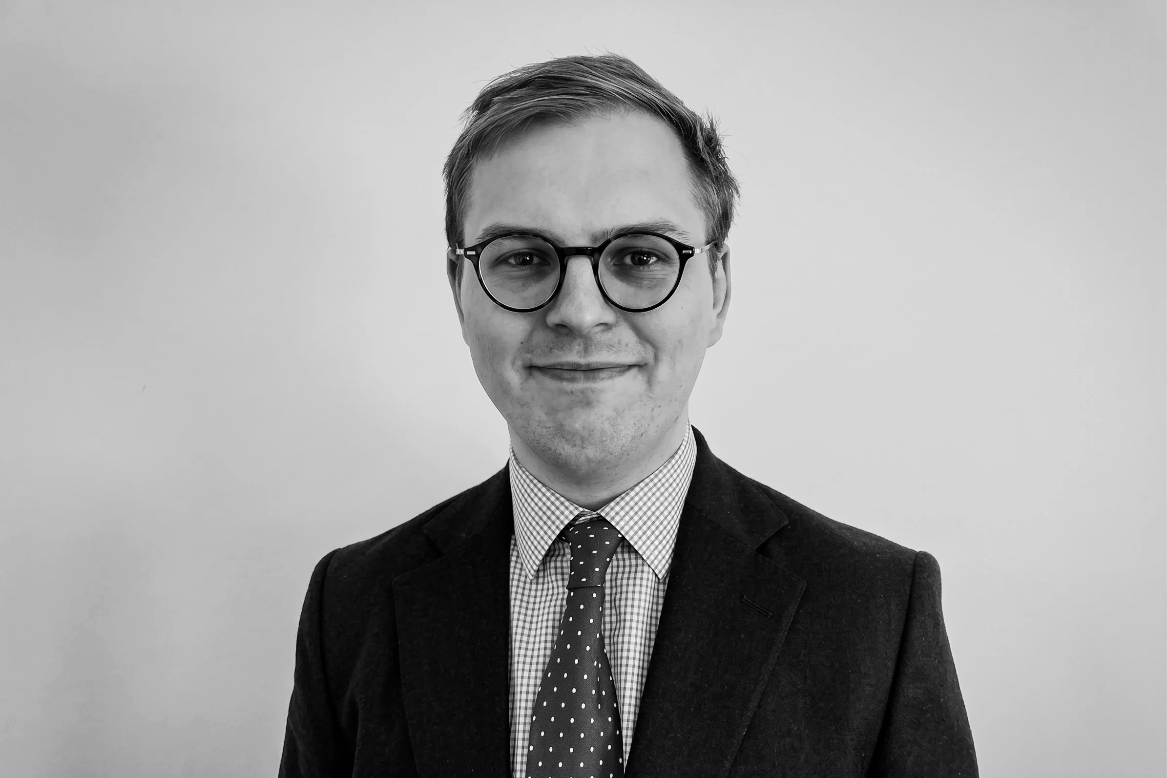 A young man with glasses, dressed in a suit with a checkered shirt and polka dot tie, smiling at the camera against a plain background.