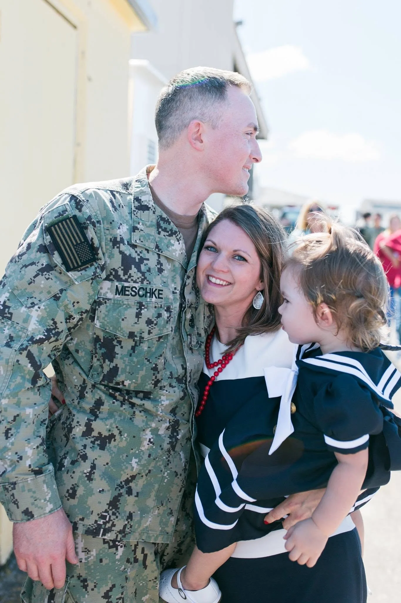 A soldier in camouflage uniform smiling, standing next to a woman and a young girl. The woman and girl are hugging the soldier; the woman is smiling, and the girl is looking at the soldier. Outdoors, with a bright sky.