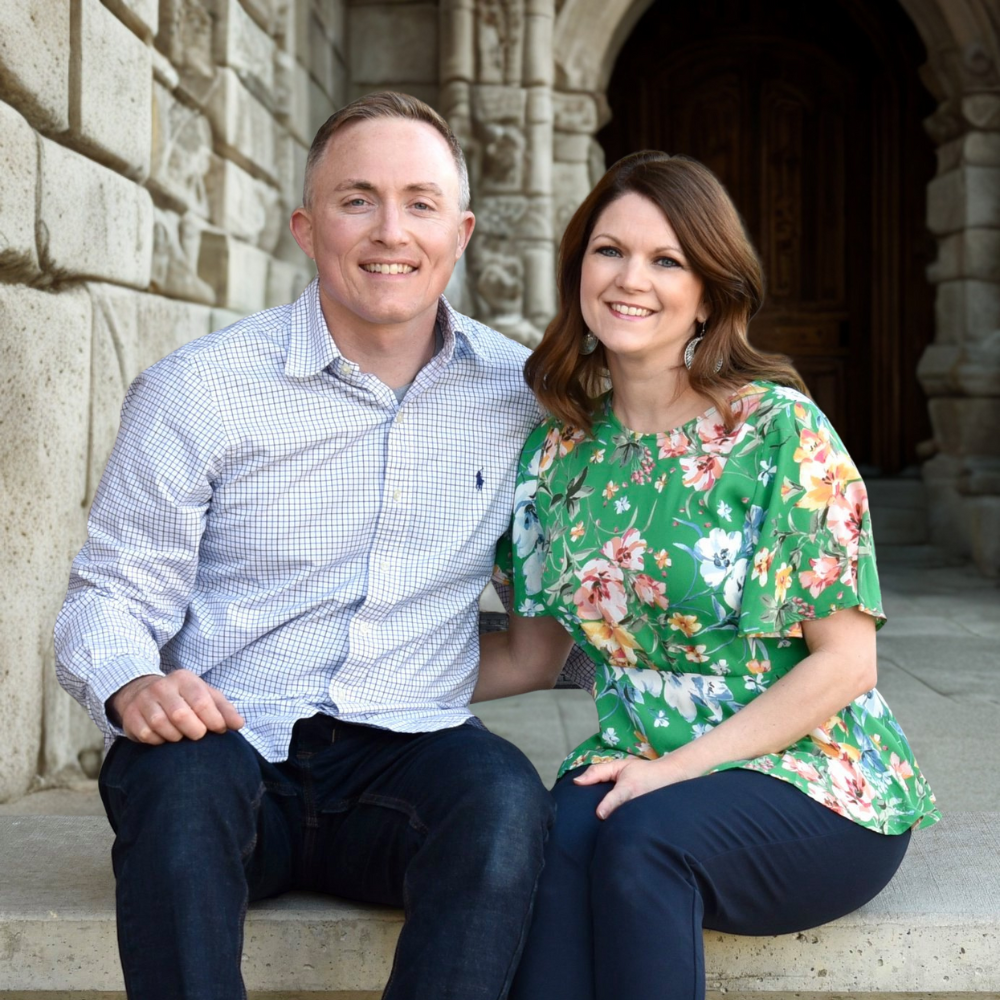 A man and woman sitting on stone steps outside a stone building, smiling at the camera.