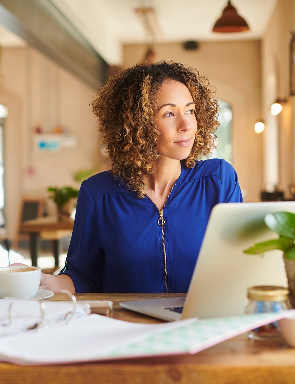 A woman with curly hair wearing a blue jacket, sitting at a wooden table with a laptop, coffee, and a plant in a cozy cafe.
