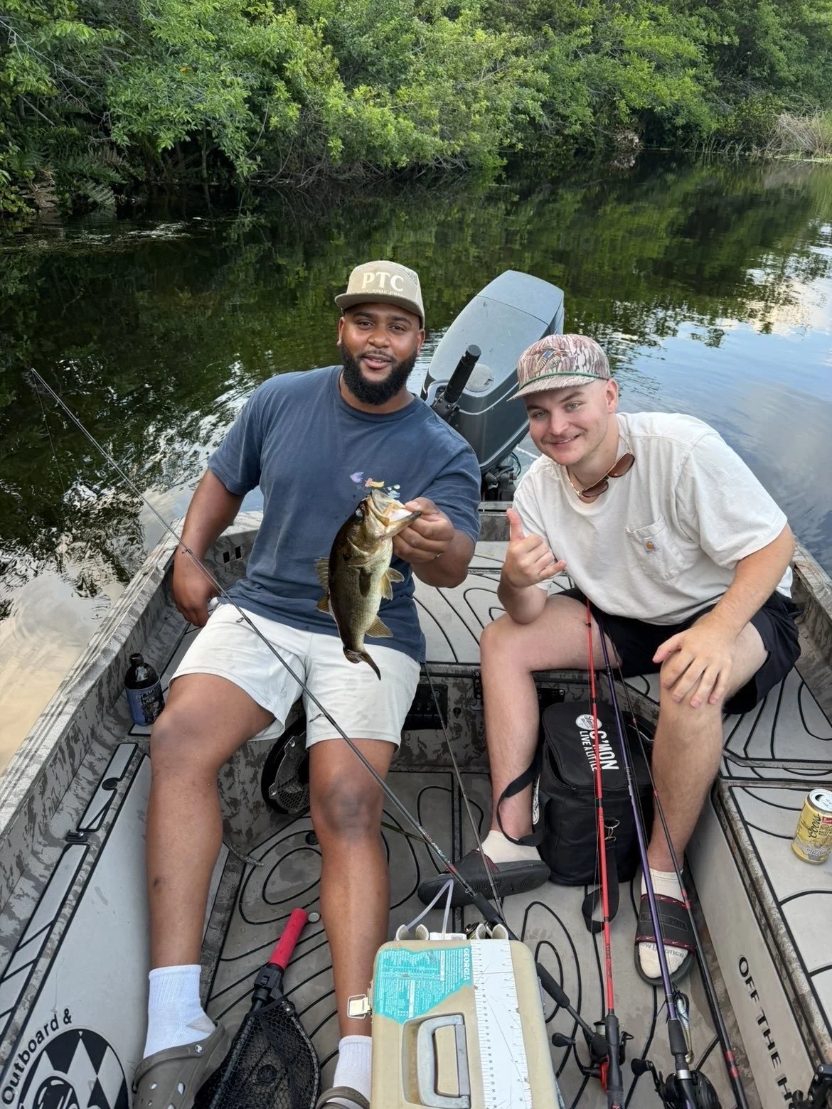 Two men on a boat on a river, one holding a large fish and both smiling at the camera, surrounded by green trees.