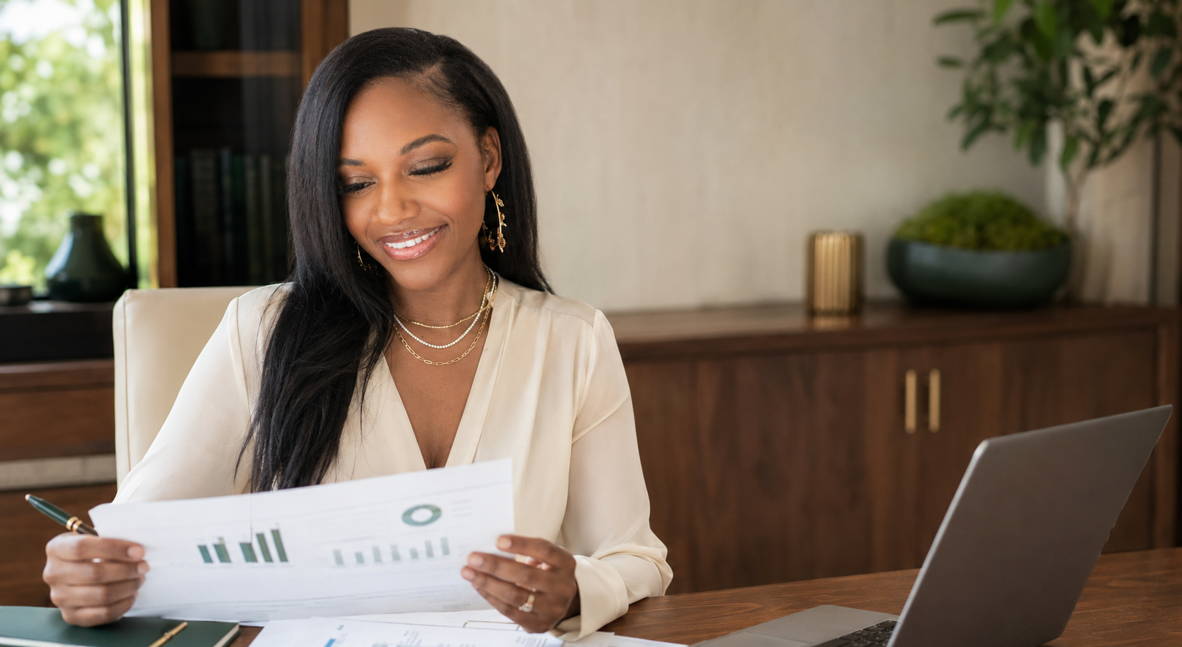 A woman with dark hair and gold jewelry smiling while reading documents at a desk with a laptop and pen, in a well-lit room with a window, plants, and wooden furniture.